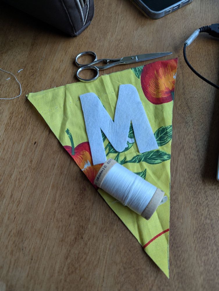 A triangular flag made of yellow cotton with an apple motif, with a white felt M placed on it. A thread reel and small scissors are next to it, on a brown table.