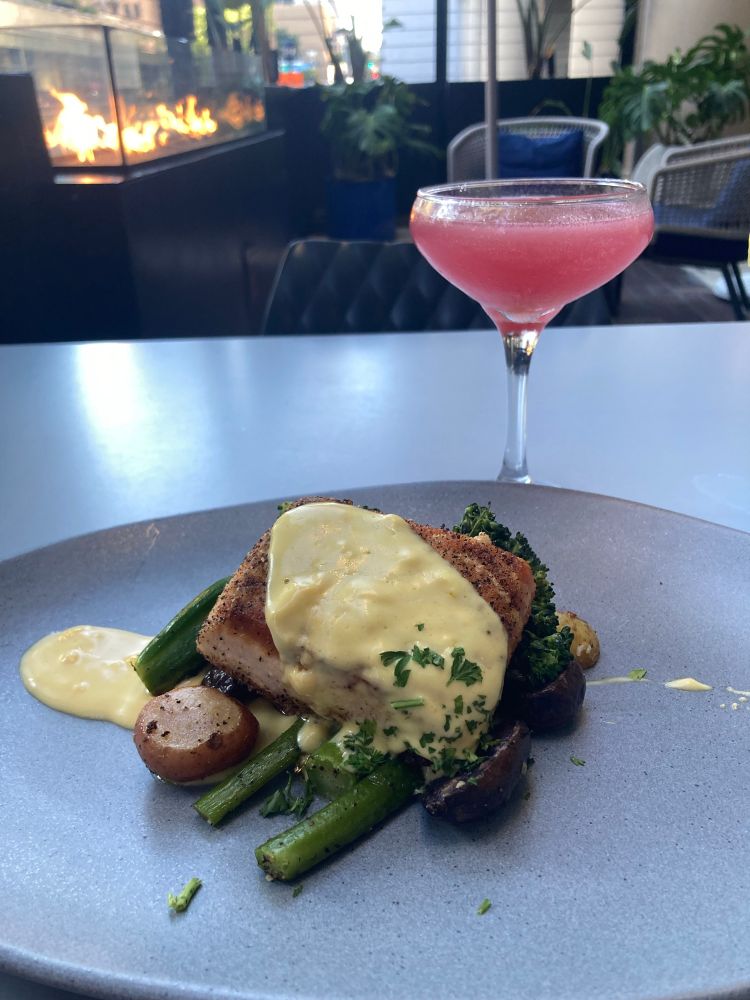 A plate of salmon dinner next to a glass with a strawberry margarita. In the background is a fire heating the hotel restaurant patio.