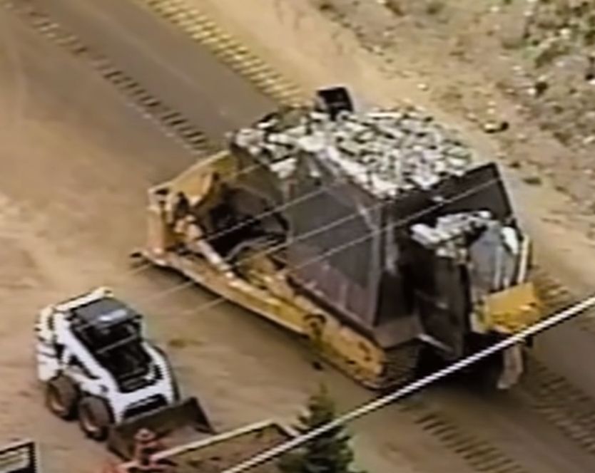 a large steel plate and concrete reinforced bulldozer drives past a parked skid loader