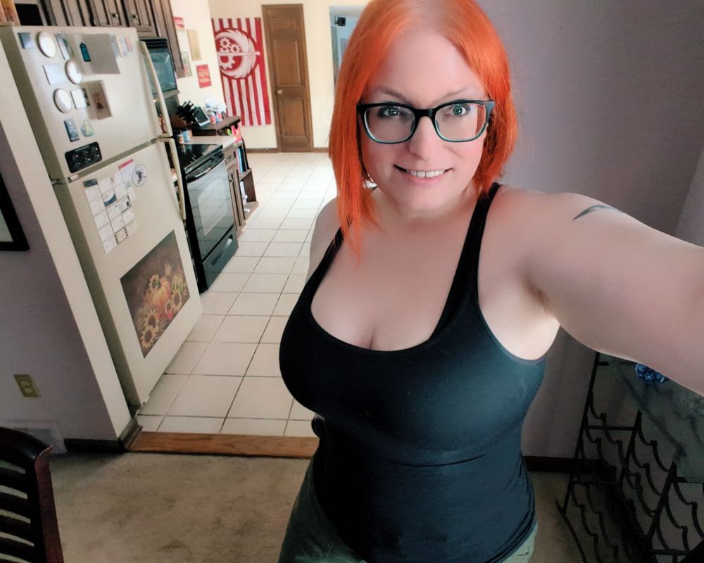 Just Jess in a tank top in her dining room with the kitchen behind. A Brotherhood of Steel flag from Fallout is on the far wall. I meant to catch some of my paintings in this pic too but you can only hold the phone so far out.