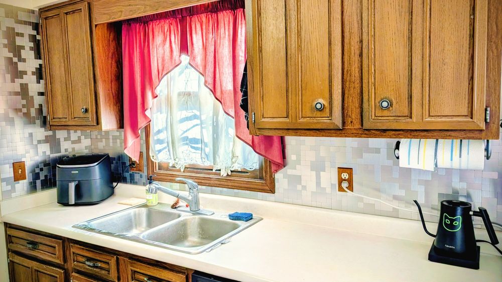 View of the sink side of kitchen and countertop. Cupboards are dark oak counter is white and the new tiles are brushed stainless