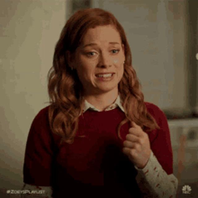 a woman with red hair is wearing a red sweater and a white shirt and is making a funny face .