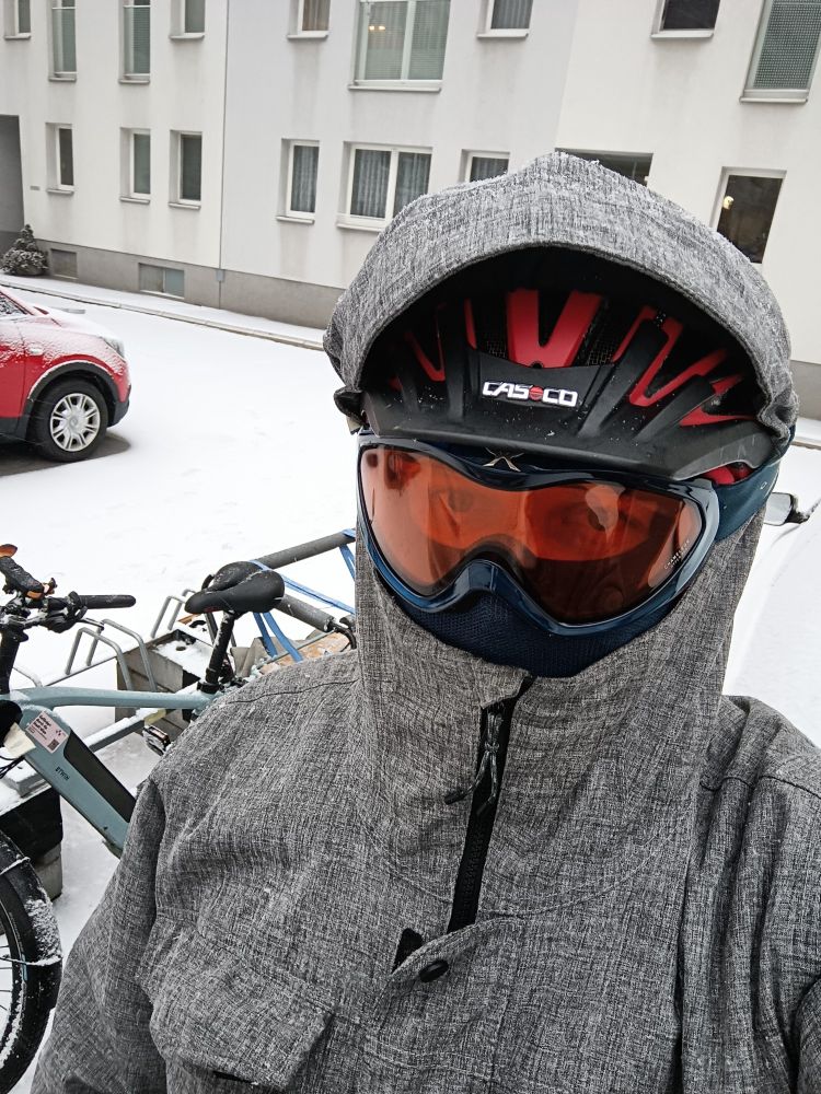 Selfie in einem Innenhof. Ein Radfahrer steht neben seinem Rad und hat einen roten Radhelm auf, darüber eine graue Winterjacke inklusive Kapuze und eine Schibrille.