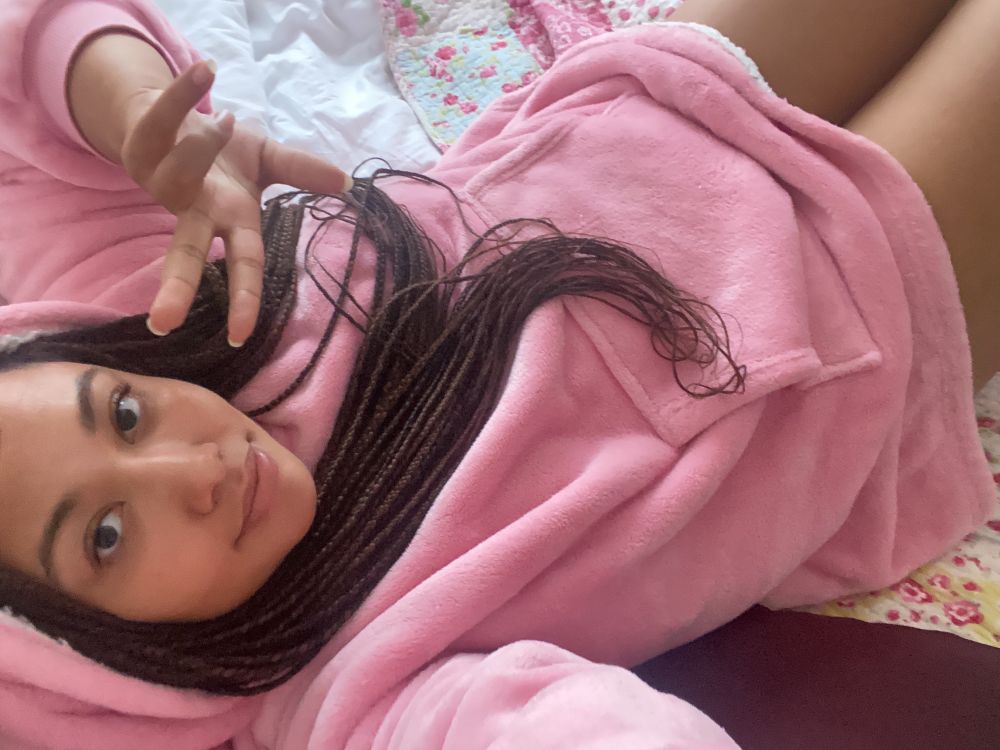Picture of me lying down in a really cozy fluffy pink hoodie. I have long braids and smiling to the camera holding two fingers up in the peace sign