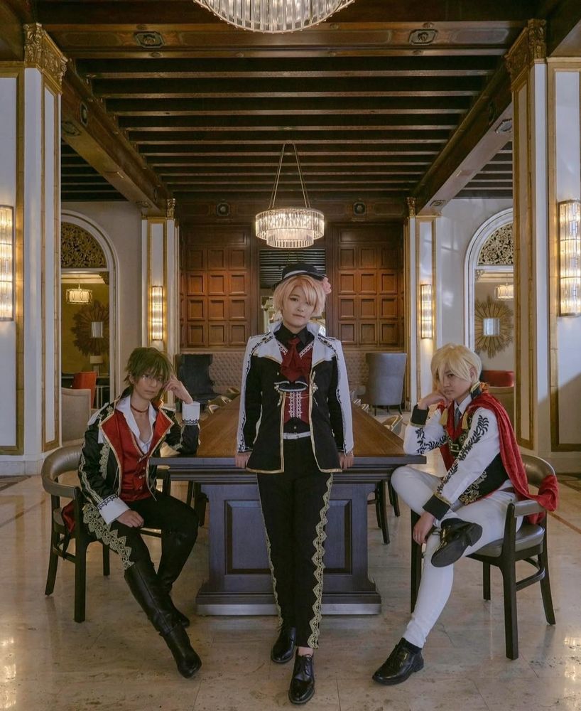 idolish7 viva fantastic life cosplay; Mitsuki in front of long table in a fancy lobby with chandelier, Yamato and Nagi on left and right side of him sitting down. 