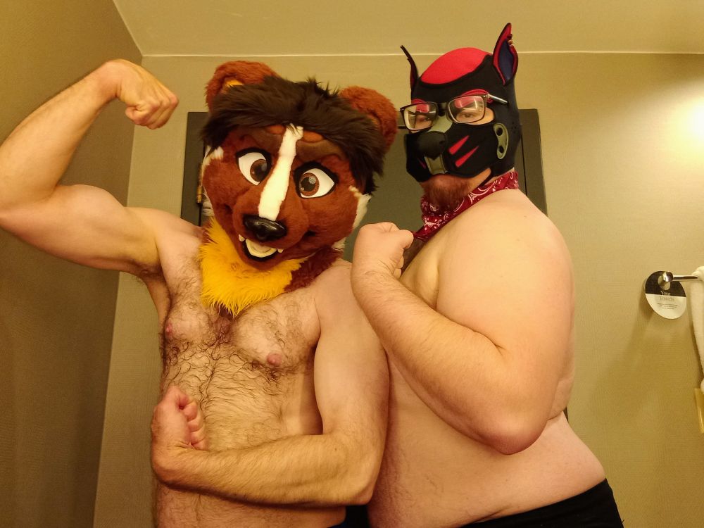 Nedsel Weasel and Pup Koda, shirtless, with fursuit head and pup hood respectively, flexing.