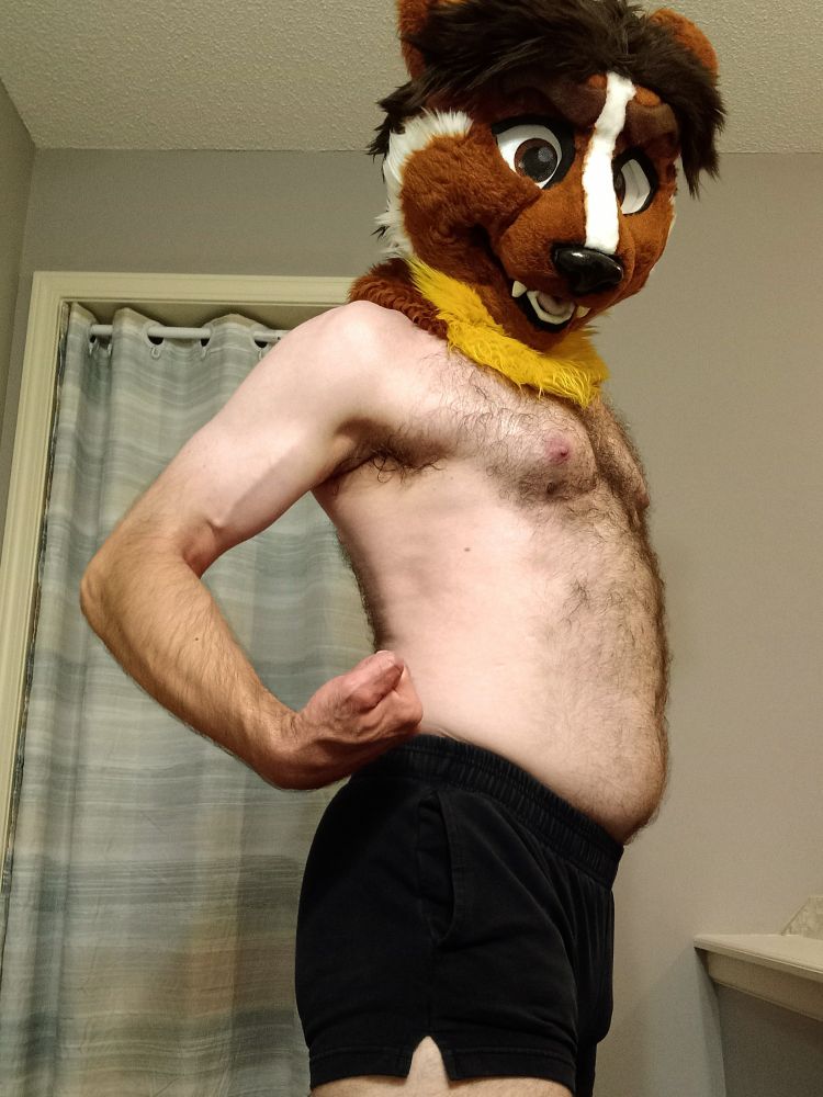 Shirtless man with weasel fursuit head, turned to the side, curling his arm at his side and puffing out his chest.