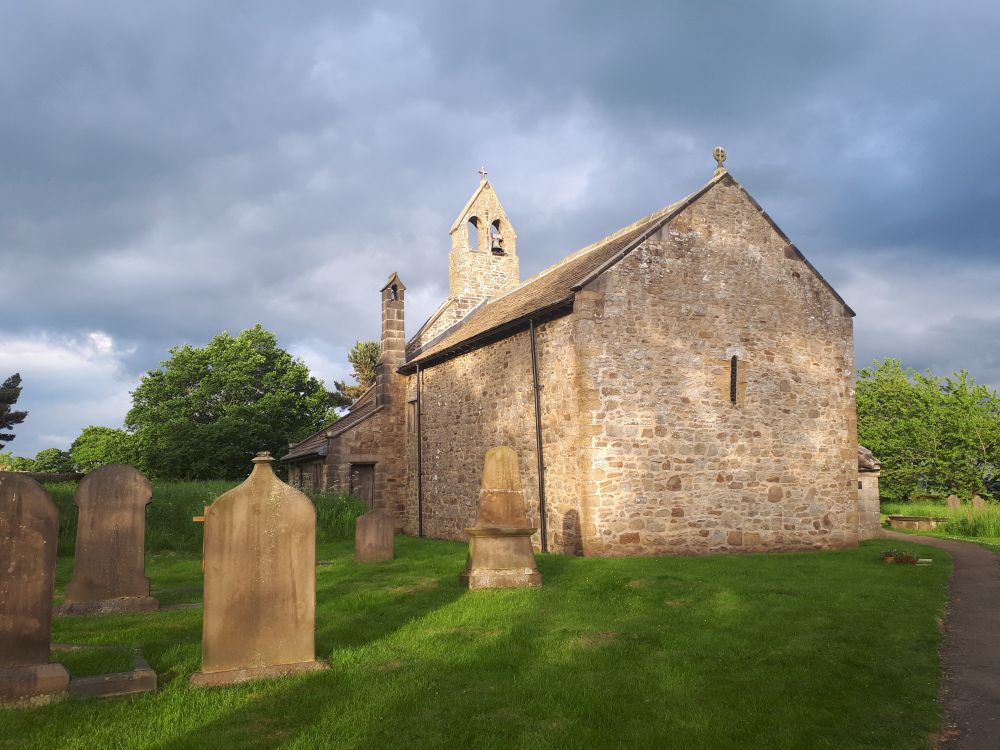 A small Norman church built of gritstone rubble, sits in a graveyard, surrounded by gravestones and trees. The bell tower contains a bell, inscribed with Latin, dated to the 14th century which reads 'John is my name'. 