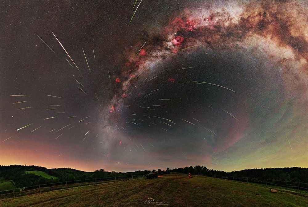 A starfield is shown above a grassy field with hills on the horizon. The band of our Milky Way Galaxy arches across toward the right. Many streaks appear emanating out from a place on the Milky Way just above the horizon. Please see the explanation for more detailed information.