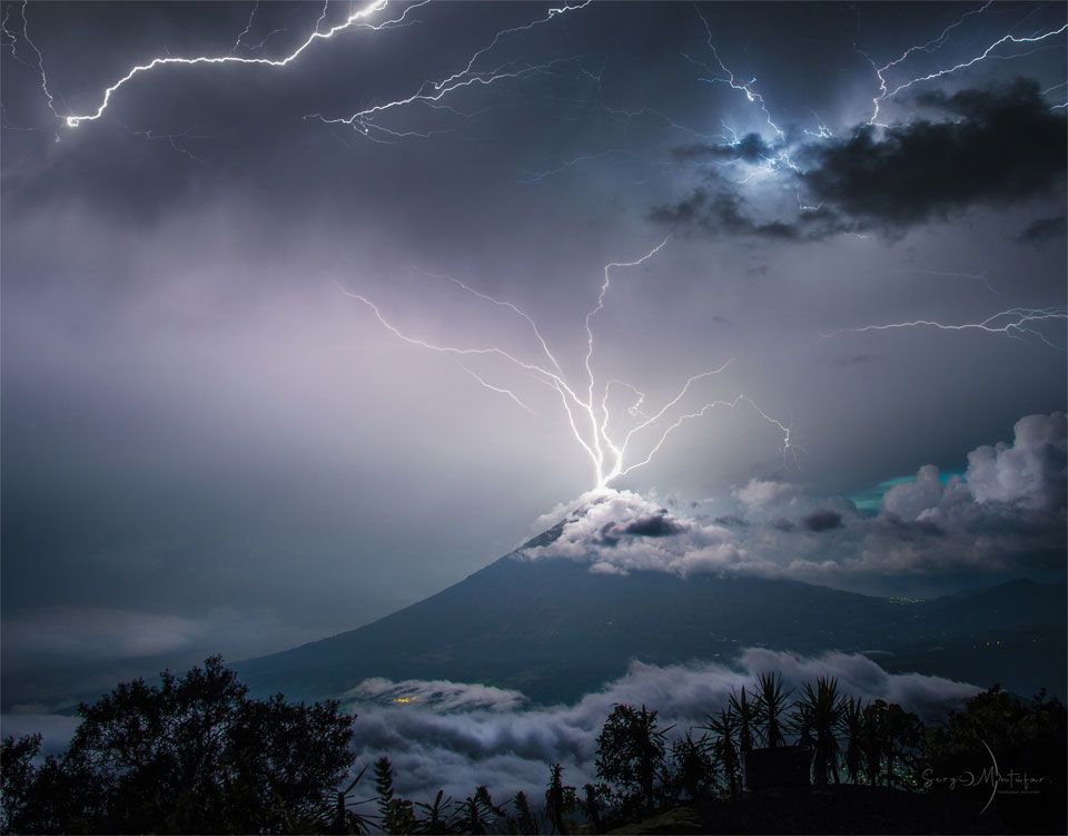 A volcano is shown with its peak in the midst of purple clouds. Lightning appears to come out of the top of the volcano in multiple paths into the upper sky. Please see the explanation for more detailed information.