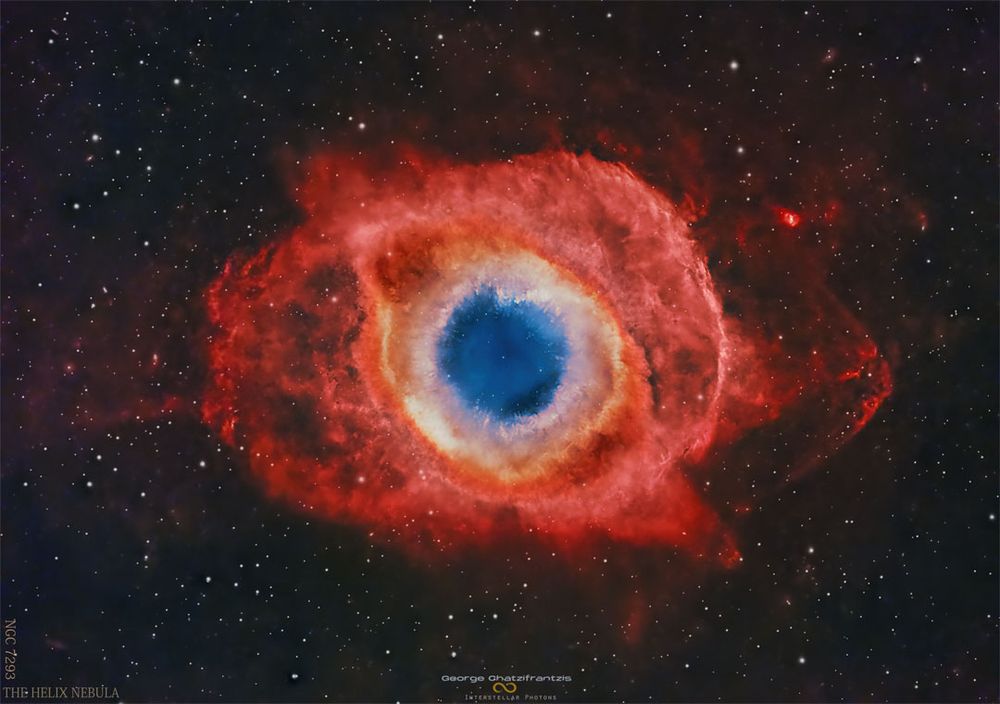A nearly circular nebula with a blue core surrounded by small white knots, an orange ring and expansive red strucures. Please see the explanation for more detailed information.