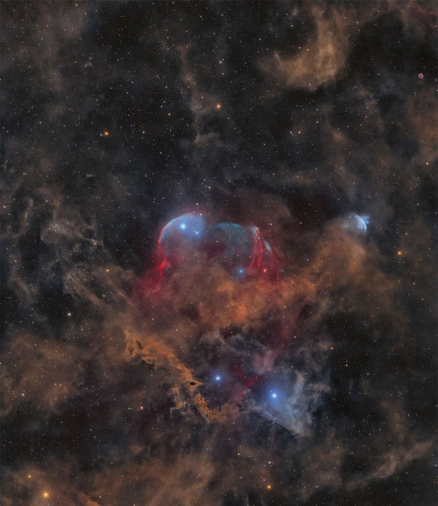 A starfield is shown with many stars and several faint light brown dust clouds. In the center is a large cloud with brown dust and gas shells lined in blue. Please see the explanation for more detailed information.