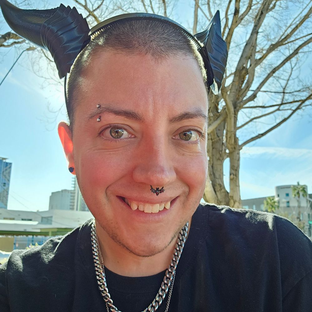 Agender smiling a fangy smile while standing in the sun and wearing black dragon horns. 