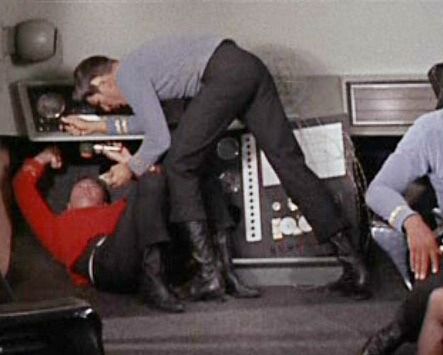Spock leaning over a panel with Scotty underneath the same panel . Spock is bent over it
