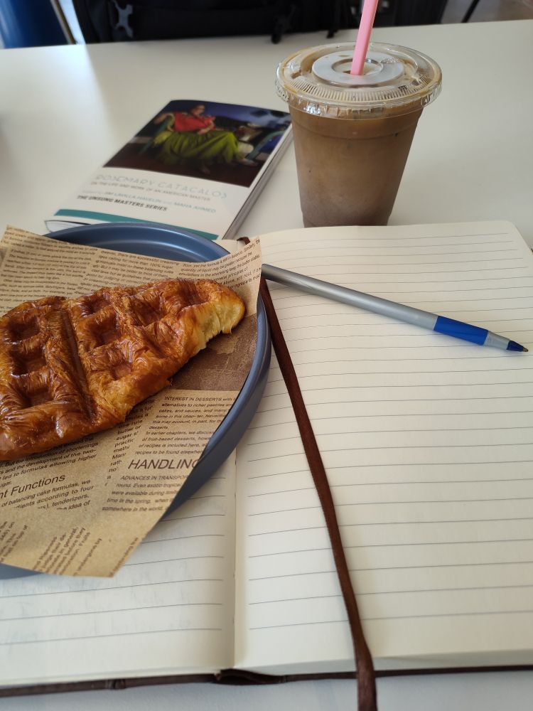 My stuff spread across a Cafe table: a journal and pen, a waffle croissant on a plate, the poetry of Rosemary Catacalos and a cookies n cream latte.