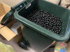 A green wheelie bin that is full of black atoms for the model sets, underneath the bin there are cardboard boxes full of grey bonds.  