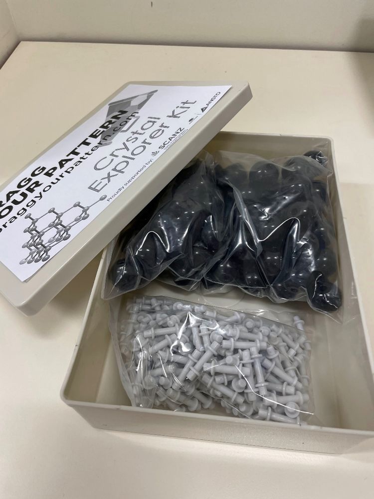image of a crystal model kit box, with the contents displayed.  Inside the box there are two packets, one of black atoms and one of grey bonds