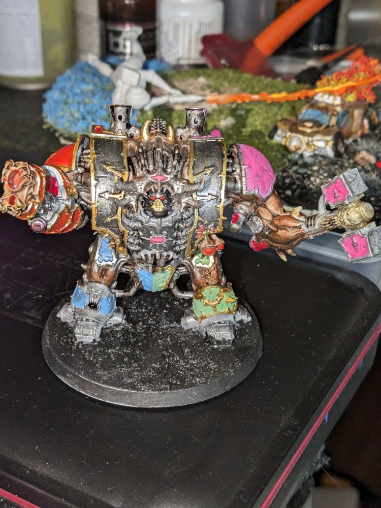 A fully painted Chaos Space Marine Helbrute sitting on a cluttered work desk. It is Voltron inspired with a black torso, red gun arm, pink hammer arm, pale blue leg, and pale green leg, with lots of gold and silver trim.