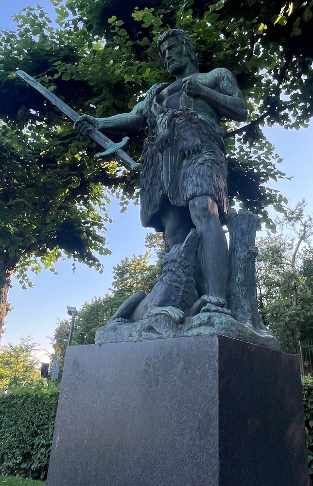 Bronze statue with green oxidation texture. Bearded man holding sword from the blade with his right hand. And statue has some animal elements too: man is wearing a wolf pelt and dog is looking up to its master laying on the ground at his feet.