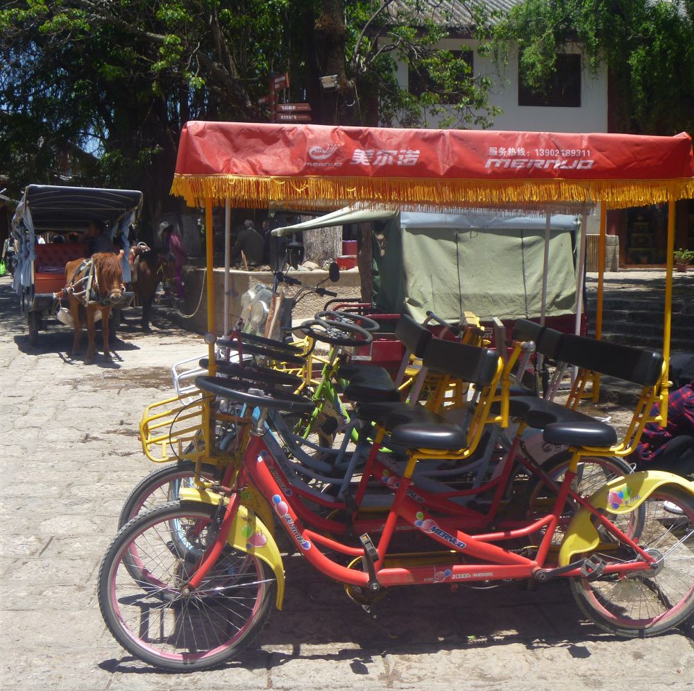 Auf einem kleinen gepflasterten Platz in der Sonne vor einer Gasse mit Gebäuden und hohen Laubbäumen stehen rechts eine Reihe rot oder grün lackierter Fahrradrikschas mit Lenkrädern, schwarzen Sätteln, gelben Schutzblechen und roten Sonnendächern. Sie tragen bunte Bubbles als Zeichnung und die Aufschriften "Amerikanisch", "Merno" und die Telefonnummer der Service-Hotline. Eine kleine Bude steht rechts dahinter mit olivgrünen Sonnenschutz-Markisen. Links am Rand steht ein Pferdefuhrwerk mit eingespanntem braunen Pferd, dahinter ist ein weiteres Pferd im Schatten angebunden. 