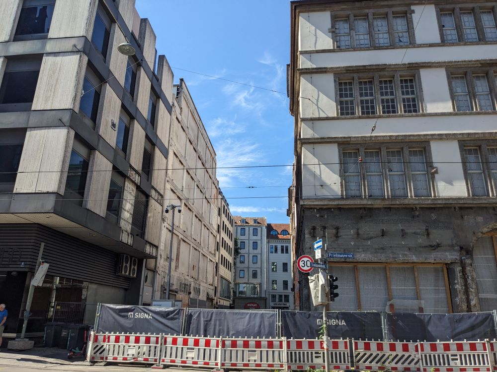 Hinter einem Absperrgitter an einer aufgelassenen Verkehrsampel mit 30er-Schild in der Prielmayerstraße, wie auf dem Straßenschild zu lesen ist, rechts ein Altbau mit drei sichtbaren Obergeschossen über dem teilabgerissenen Erdgeschoss. Die Fenster haben je 8 Teilstücke mit je 9 kleinen Scheiben. Zwischen den Geschossen ein glattes helles Band mit Steinumrandungen wie an den Fenstern. Im Durchblick zwischen den Gebäuden erḱennt man die mehrstöckigen Gebäude der Parallelstraße. Ein Teil der Front des Neubaus links ist provisorisch verschlossen. Der Neubau selbst hat senkrechte helle Beton-Bänder gegenüber waagerechten dunklen Metallbändern an der Außenfassade, zwischen denen dunkle einteilige Fenster liegen. Verbarrikadierte Erdgeschosse, Planen und unbekleidete Fassaden, an denen Trambahnen vorbeifahren.