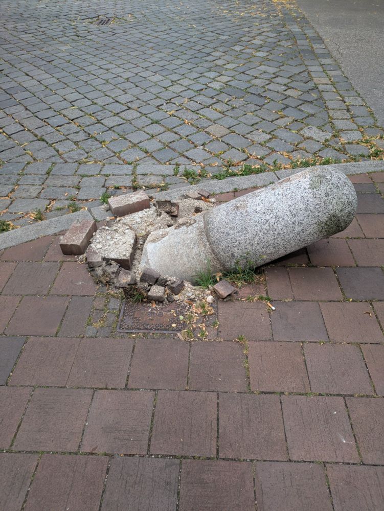 An einem Straßeneck mit Gehweg aus roten Platten und Kreuzung aus Pflastersteinen liegt ein Poller aus Granit umgestoßen auf dem Gehweg. Man sieht, dass er zu einem Drittel eingeraben war. Beim Umstoßen sind Mörtel und Einsatzsteine herausgerissenworden und liegen herum.