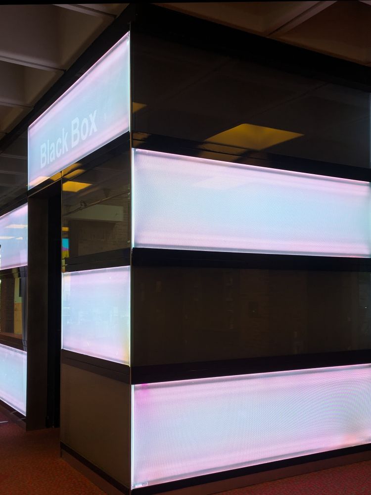 Innenaufnahme eines Eingangs zu einem Veranstaltungsort, der mit schwarzen und weißen Blenden mit Hintergrundbeleuchtung dekoriert ist. Der Text Black Box ist links oben als Leuchtschrift zu lesen.