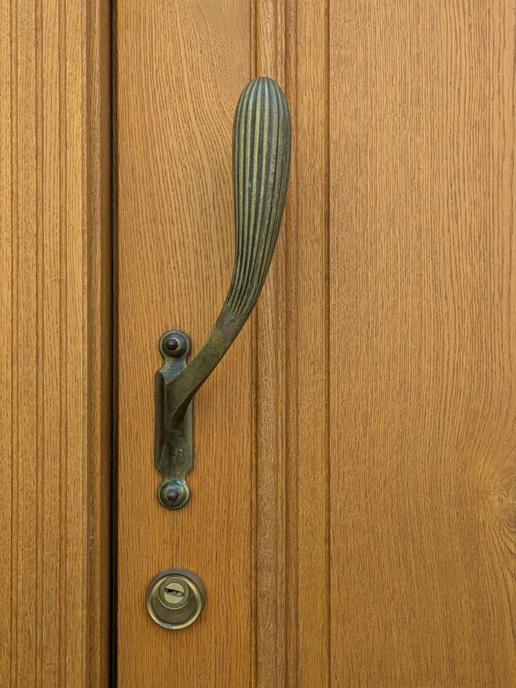 Ausschnitt aus einer hellen Holztür mit Rahmen, wohl aus Eichenholz. Unten ist der Zugang zum Sicherheitsschloss in einem runden Messingeinsatz, daüber ist senkrecht ein Griff aus Metall mit Patina angebracht, der wie eine geriffelte Keule nach oben gebogen ist.