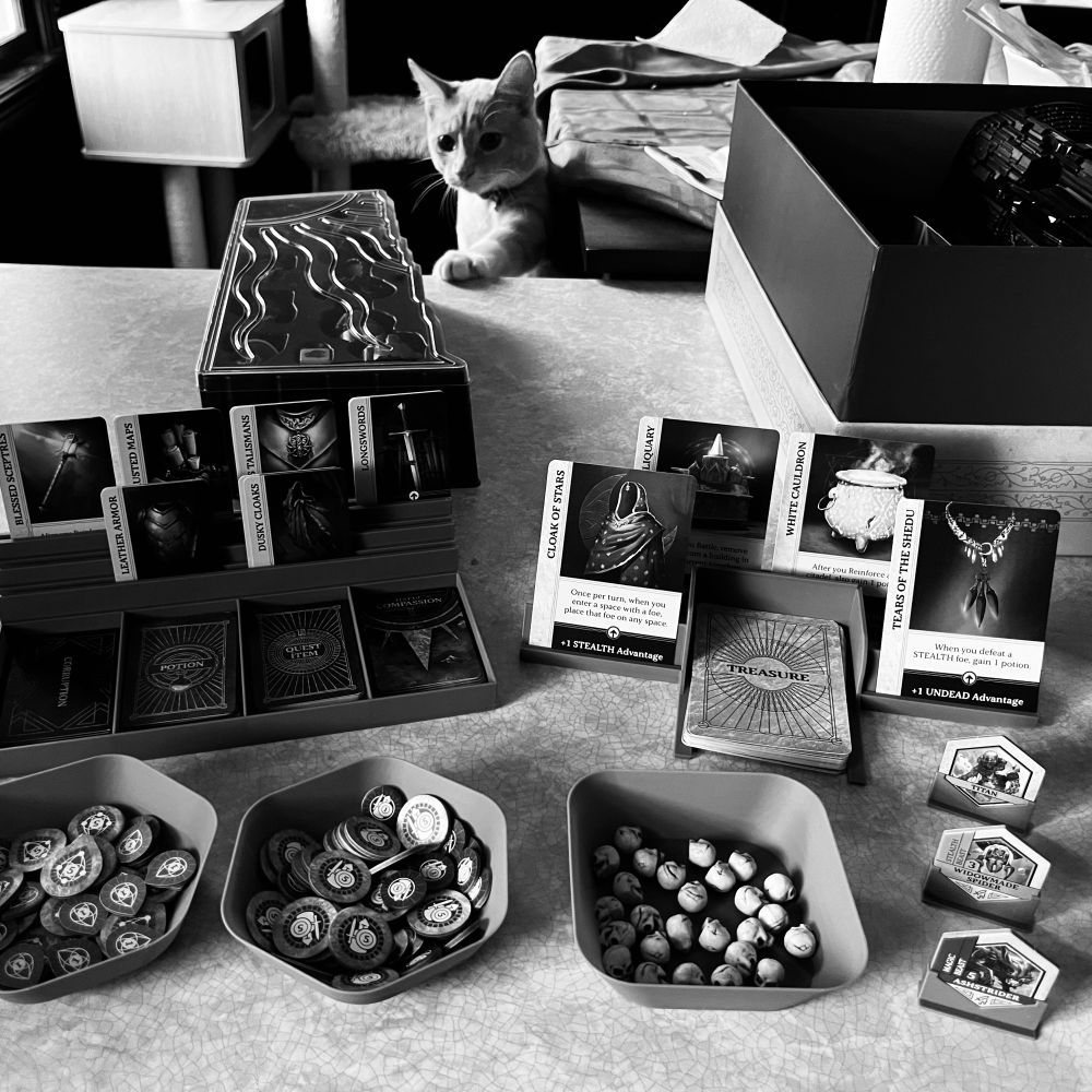 Components for the board game, Return to Dark Tower, laid out on a countertop. Several 3D printed trays hold the card decks, game tokens and skulls. Yes, skulls. Lucky Leo, a cream and white kitten, is inspecting the scene in the background.