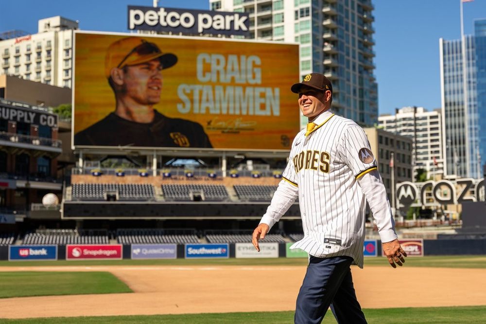Photo of Craig Stammen smiling and walking on the field at Petco Park. He is wearing a brown Padres hat and a home white pinstripe jersey. The board behind him has a gold graphic that reads "Craig Stammen | Manager".