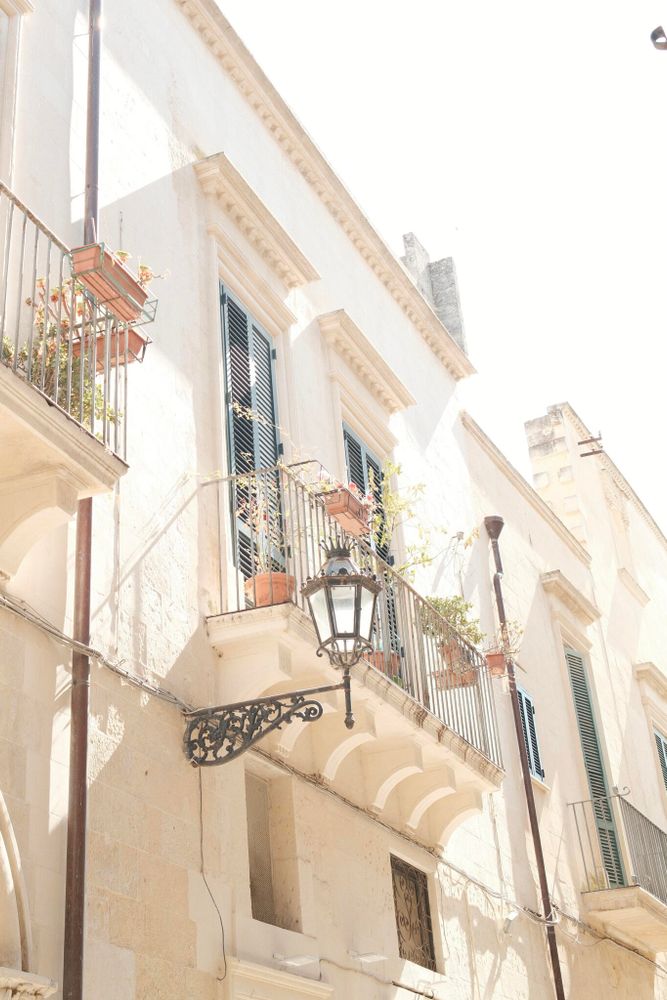 A sunlit street scene featuring a white, historical building with green shutters. The building has small balconies adorned with potted plants. An ornate metal bracket holds a vintage lantern. Sunlight casts soft shadows against the building's facade.