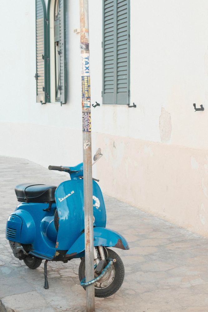 A vintage blue Vespa scooter is parked on a paved sidewalk next to a white building with green shuttered windows. The scooter, showing signs of wear, is locked to a pole with various stickers on it. The sun casts soft light on the scene, creating a tranquil atmosphere.