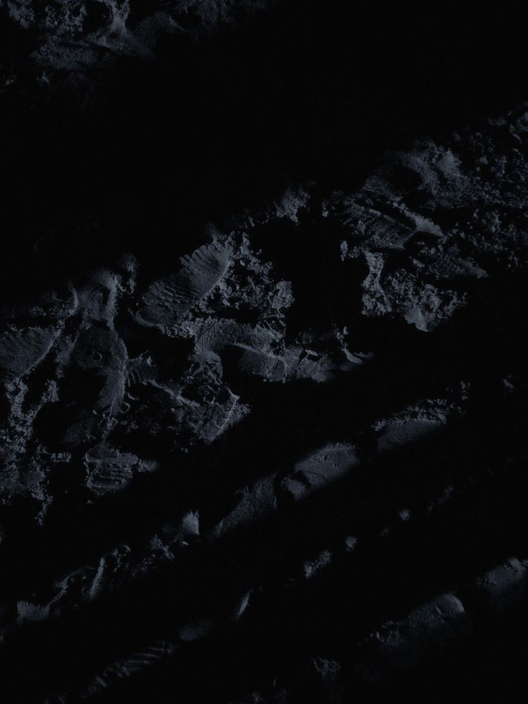 Dark, shadowy image of textured ground, possibly with footprints or patterns. The surface appears uneven and covered with fine details, illuminated by minimal light with deep shadows.