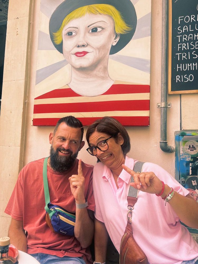 A man and woman are posing playfully in front of a painting outside. The painting features a person wearing a black hat and a red and white striped shirt. The man is wearing a red shirt and has a multi-colored fanny pack, while the woman is in a pink shirt with glasses and a brown crossbody bag. Both are pointing upwards with their index fingers. There are parts of other paintings and a menu board visible to the right.
