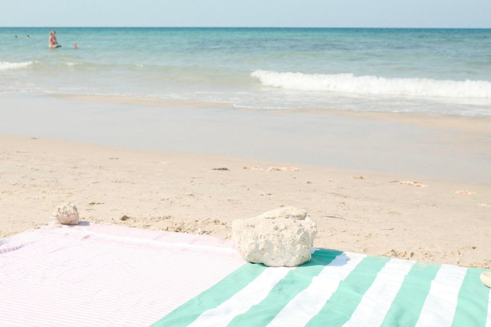 A sandy beach with two striped towels laid out on the sand, one pink and white, the other green and white. Each towel has a small rock placed on it to keep it in place. The ocean waves gently lap against the shore in the background, with a few people swimming in the distance under a clear blue sky.
