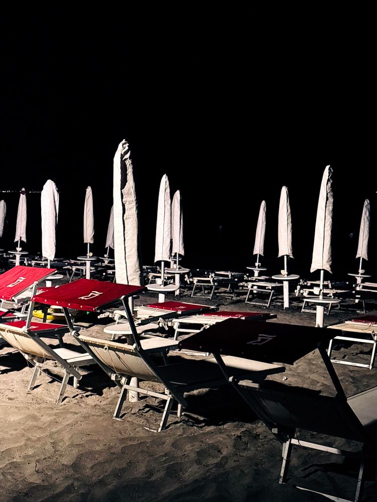A nighttime beach scene featuring rows of red lounge chairs lined up on the sand, with numerous closed white umbrellas standing upright. The dark sky and lack of people create a tranquil and deserted atmosphere.