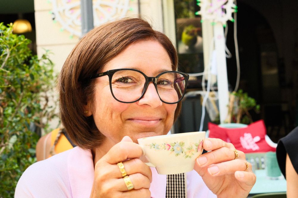 A woman with short brown hair and glasses is smiling while holding a floral-patterned teacup. She is outdoors, seated in an area with potted plants and festive decorations in the background. She wears a light-colored top and has gold rings on her fingers.