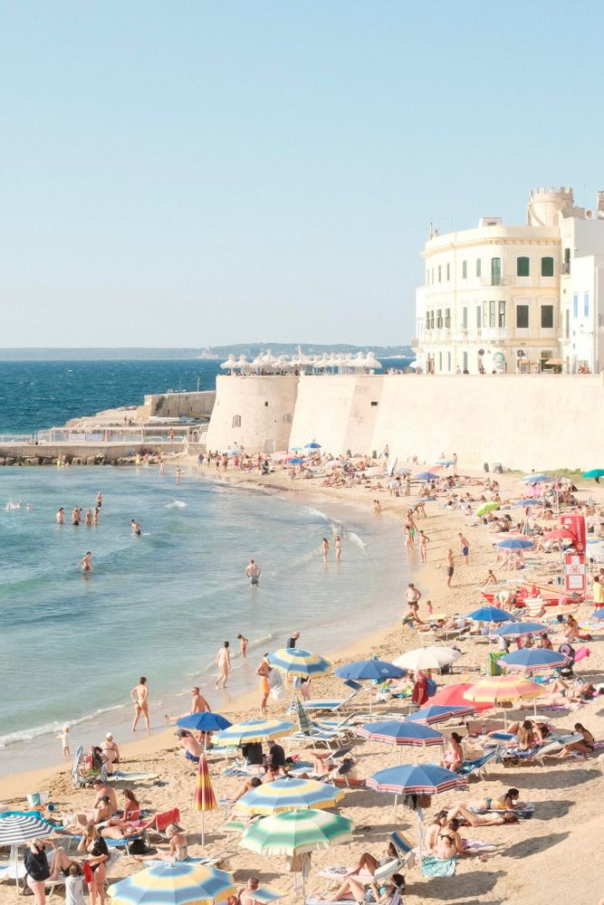 A sunny beach scene featuring many people sunbathing and swimming. Colorful umbrellas and beach towels are scattered along the sandy shore, and individuals are seen enjoying the water. In the background, there are historical buildings with a light facade overlooking the beach and the blue sea. The sky is clear, enhancing the bright and lively atmosphere.