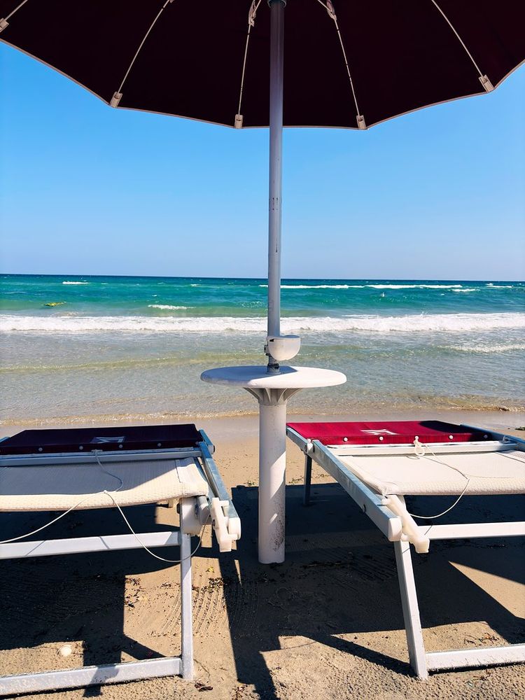 Beach scene featuring two lounge chairs under a large umbrella facing the ocean. The chairs have red cushions, and the sandy shoreline leads to gentle waves under a clear blue sky.