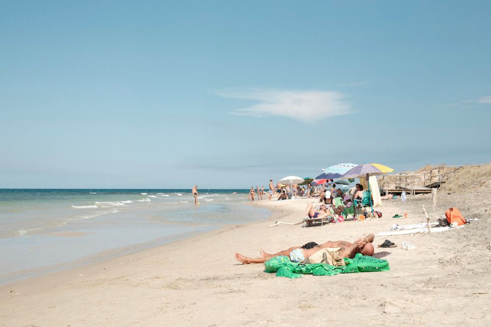 A sandy beach scene with a clear blue sky, featuring several people relaxing. In the foreground, a person is lying on a green towel. Further along the beach, other groups are sitting under colorful umbrellas near the water's edge. A few people are walking along the shoreline. The ocean is calm, and the landscape includes sandy dunes with wooden fencing in the background.