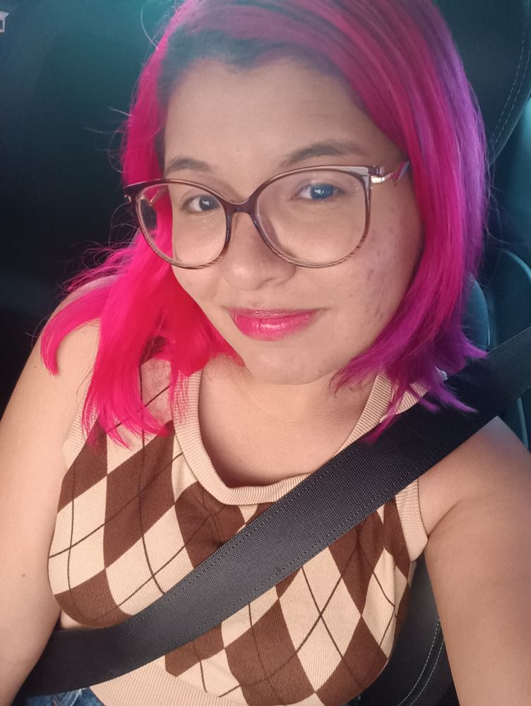 Menina normal jovem, de óculos, cabelo rosa e roxo, num carro com o cinto de segurança. Camisa quadriculada e um sorriso singelo de boca fechada.