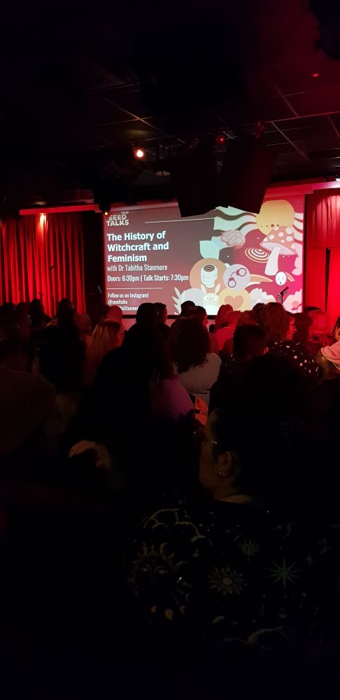 A theatre type space (actually the glee club) a busy silhouetted crowd. Waiting for the talk to start. Facing red curtains that are lit and open to reveal a screen that says The History of Witchcraft and Feminism on left side and cartoon images of witches hats, mushrooms and more that I can't make out! 