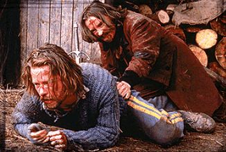 Two bedraggled, long-haired, white-skinned men on the straw-covered ground. One, in a blue knit sweater and yellow-striped uniform trousers of the Mexican-American War era, crawls away from the other, who wears a red overcoat, has a bloody cross daubed on his forehead, and is trying to pull the first one back towards him. The fashionably ornate curved hilt and grip of a _dagger_ extends from the blue-sweater'd man's back, and the wood-stave wall and stacked firewood in the background suggest a rustic environment.