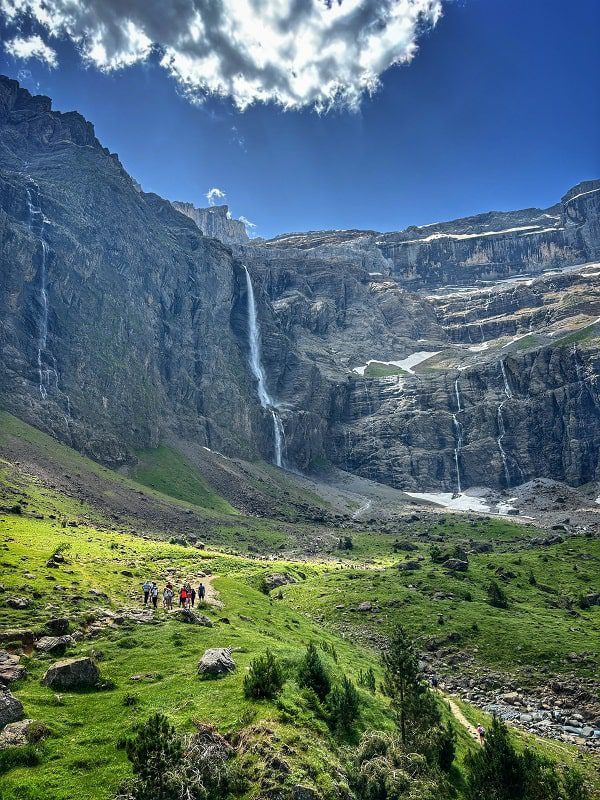 Photo of the Cirque de Gavarnie, which are tall cliffs in the mountains where multiple waterfalls cascade down the rock. It’s a bright sunny day and the vegetation around the cliffs is incredibly green. For the sense of scale, hikers are on the photo and they appear completely minuscule compared to the sheer size of the cliffs