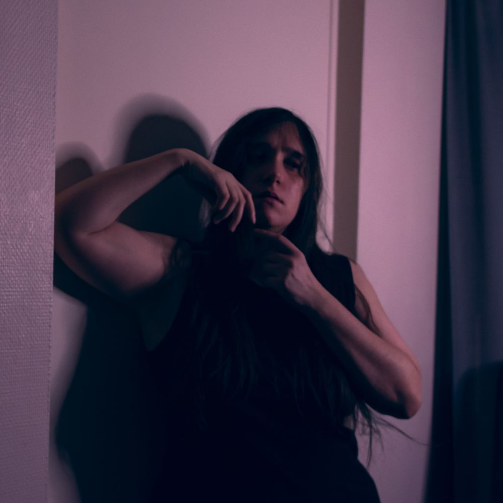 Une personne blanche avec des cheveux bruns, posant dans l'encadrement d'une porte avec les mains nouées au niveau du visage