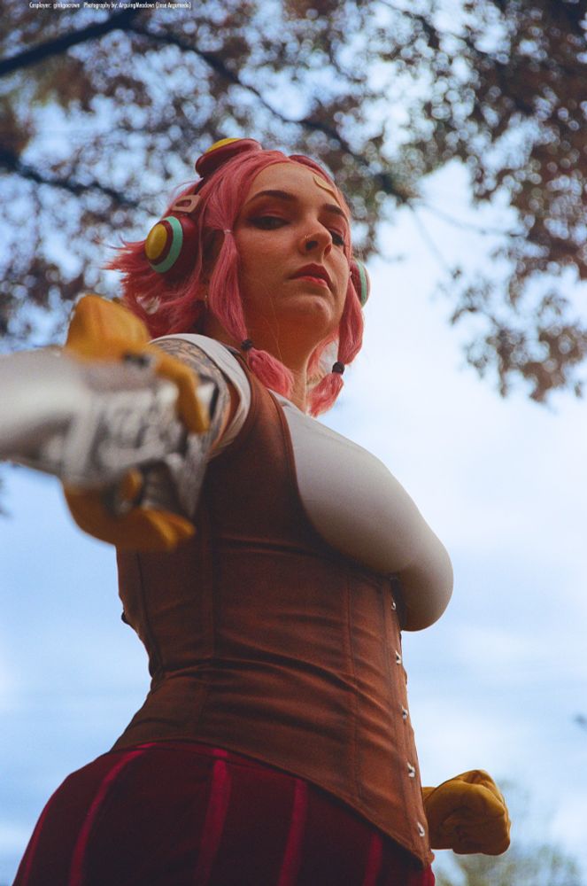A photograph of a Ginny cosplayer. She looks down the barrel of her gun at the camera and glares. 
