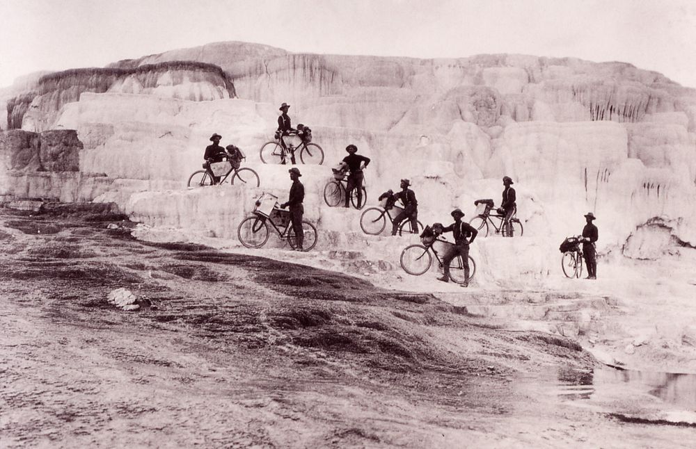 Eight Buffalo Soldiers, the first professional peacetime Black volunteer U.S. Army recruits, stand on the terraces with bicycles at Mammoth Hot Springs in Yellowstone National Park. The historic photo from 1896 is in black and white.