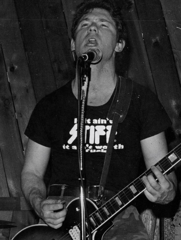 Black and white image of a guitarist playing an imitation Les Paul and attempting to sing. He wears a black t-shirt emblazoned with the phrase, "If it ain't STIFF, it ain't worth a fuck," a promotional item from UK-based Stiff Records.