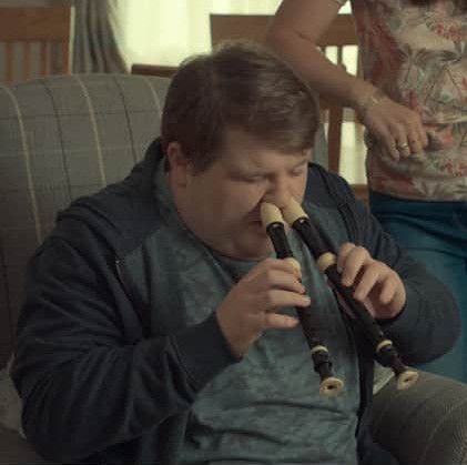 A white man is sitting in a chair. He has a recorder in each hand, and each recorder is being played in one of his nostrils. The image is a screenshot from the show "After Life" (season 1, episode 2).