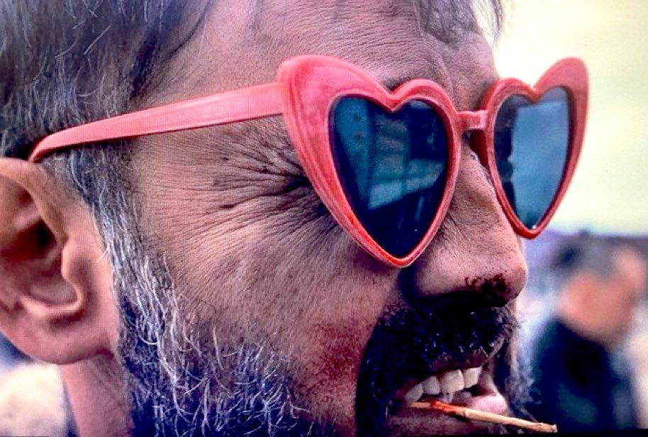 Steppenwolf still - closeup up man’s face wearing heart shaped sun glasses, he has a peppered beard and is making a grimace face while bloody teeth exposed while chewing on tooth pick
